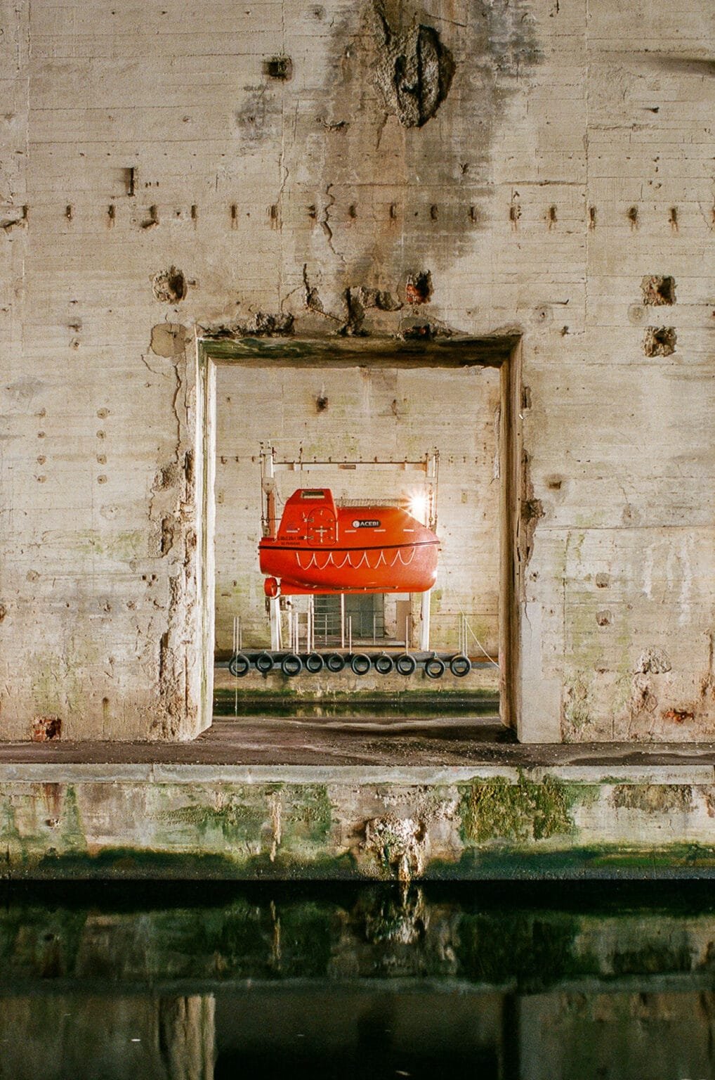 Canot de sauvetage rouge suspendu à la base sous-marine de Saint-Nazaire, encadré par un mur en béton brut.