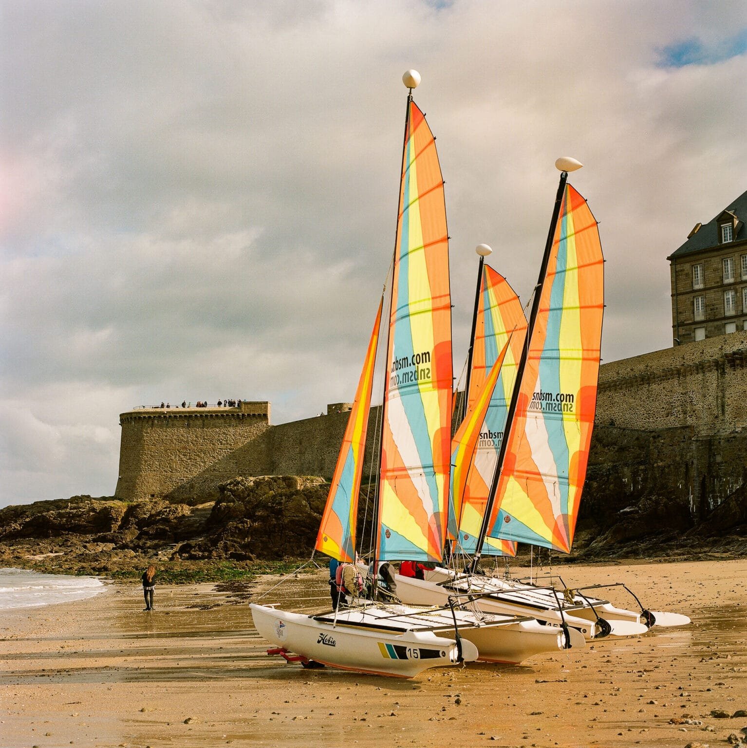 Trois catamarans Hobie Cat alignés sur la plage de Saint-Malo, avec leurs voiles colorées déployées et les remparts de la ville en arrière-plan.