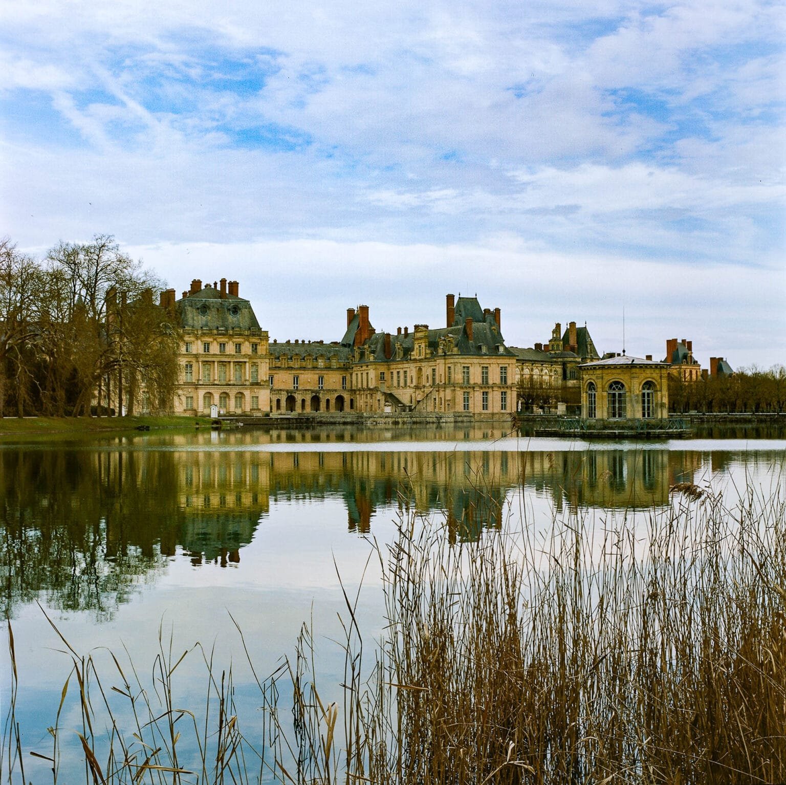 Vue du Château de Fontainebleau se reflétant sur le plan d’eau, sous un ciel légèrement nuageux.