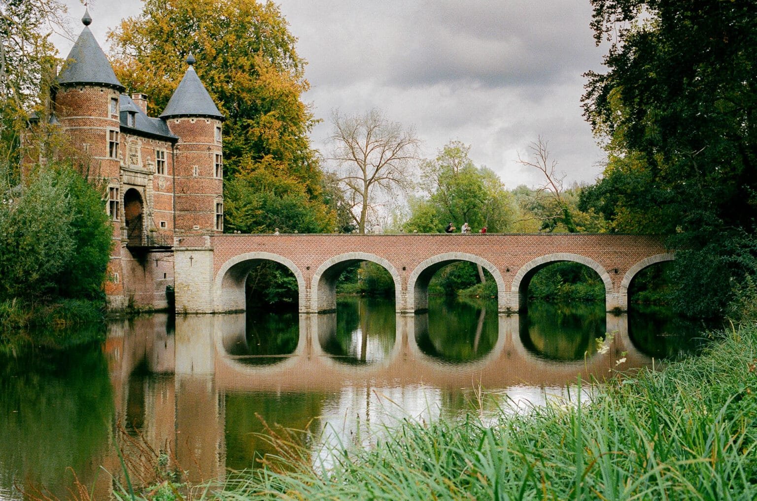 Château de Grand-Bigard en Belgique, vu depuis le pont en brique rouge, entouré d’arbres aux couleurs d’automne, reflet sur un étang calme.
