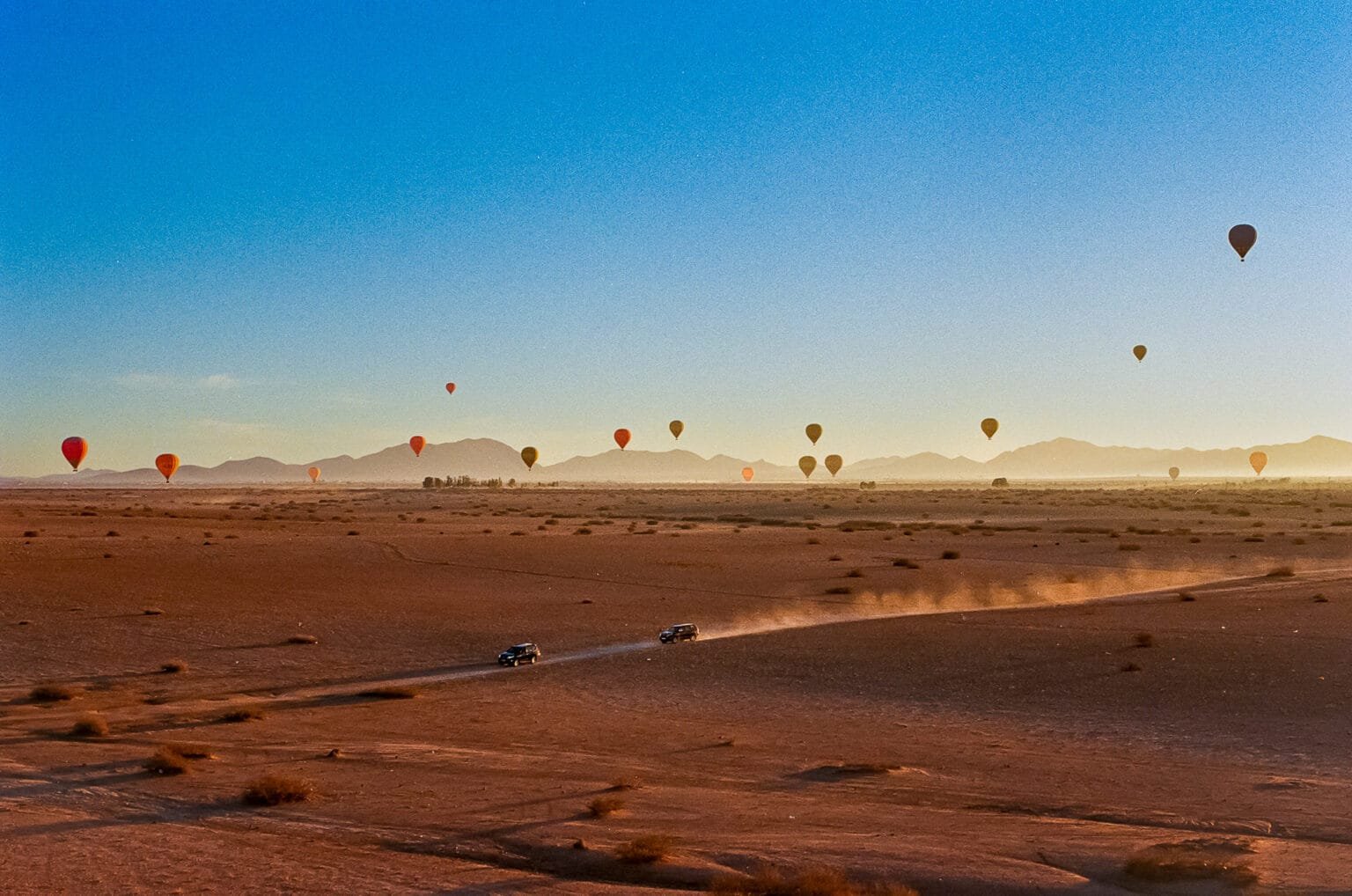 Vue panoramique sur le désert d’Agafay au Maroc avec des montgolfières en vol et deux 4x4 soulevant la poussière sur une piste.