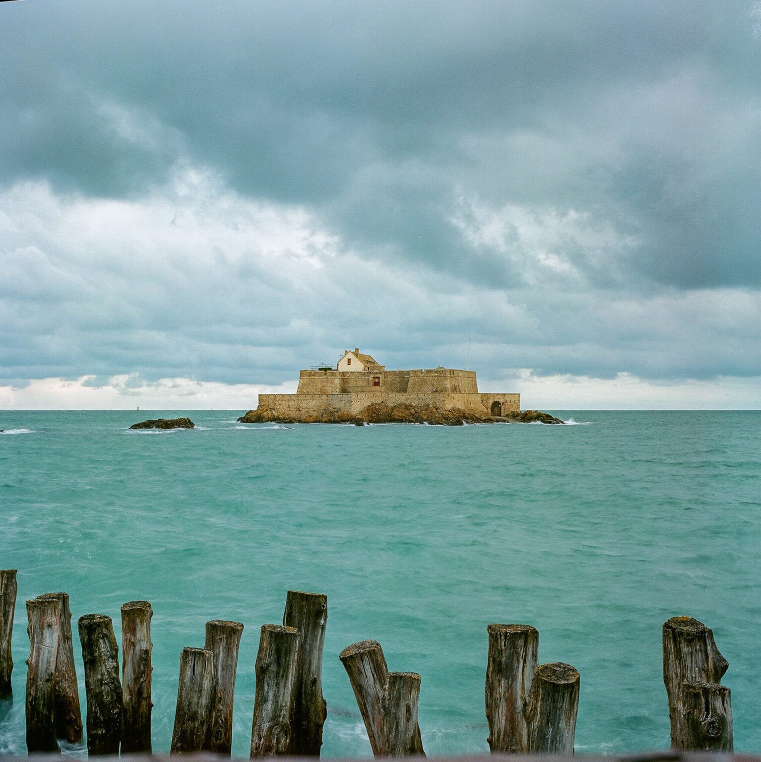 Vue sur le Fort National de Saint-Malo, entouré de l’océan vert émeraude sous un ciel orageux.