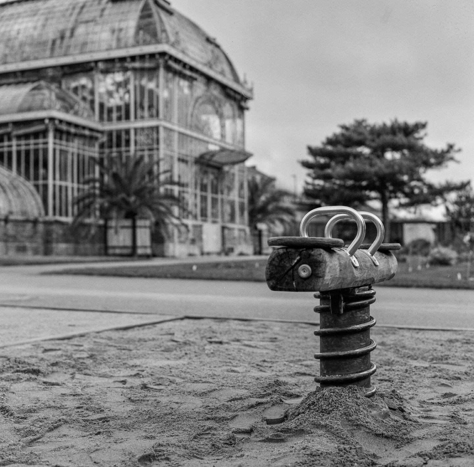 Jeu à ressort en bois sur une aire de jeux, devant la grande serre du Jardin des Plantes de Nantes, photographié en noir et blanc avec un Hasselblad 500 C/M.