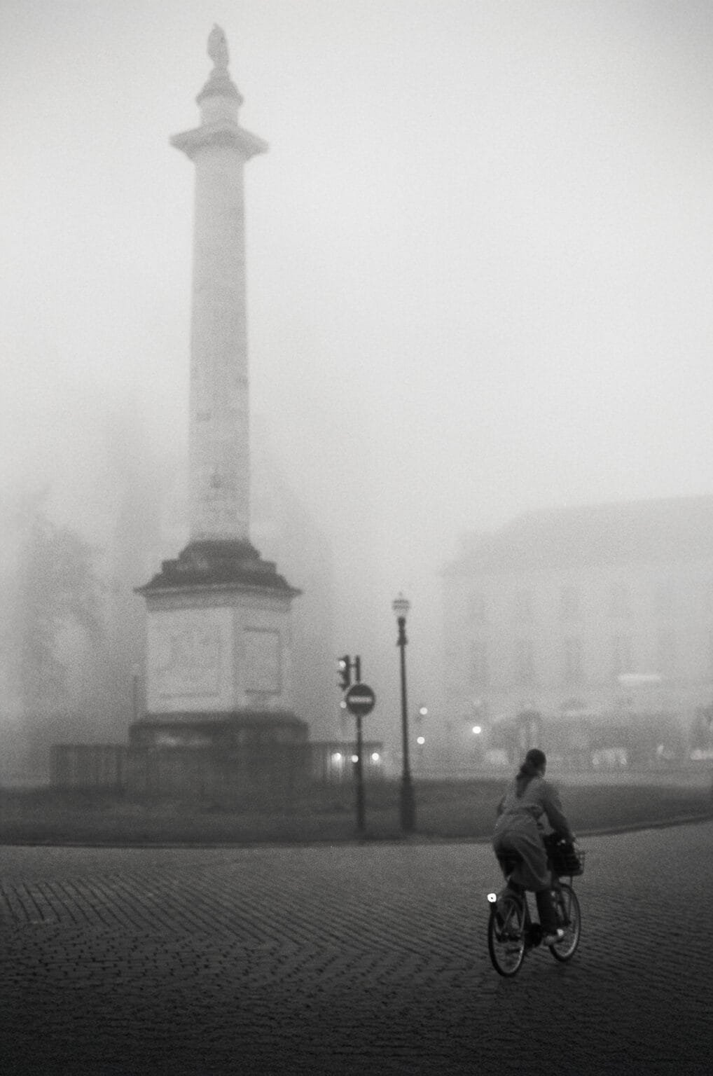 Cycliste traversant la place Maréchal Foch à Nantes, enveloppée dans une brume dense, avec la colonne surmontée de la statue de Louis XVI en arrière-plan.