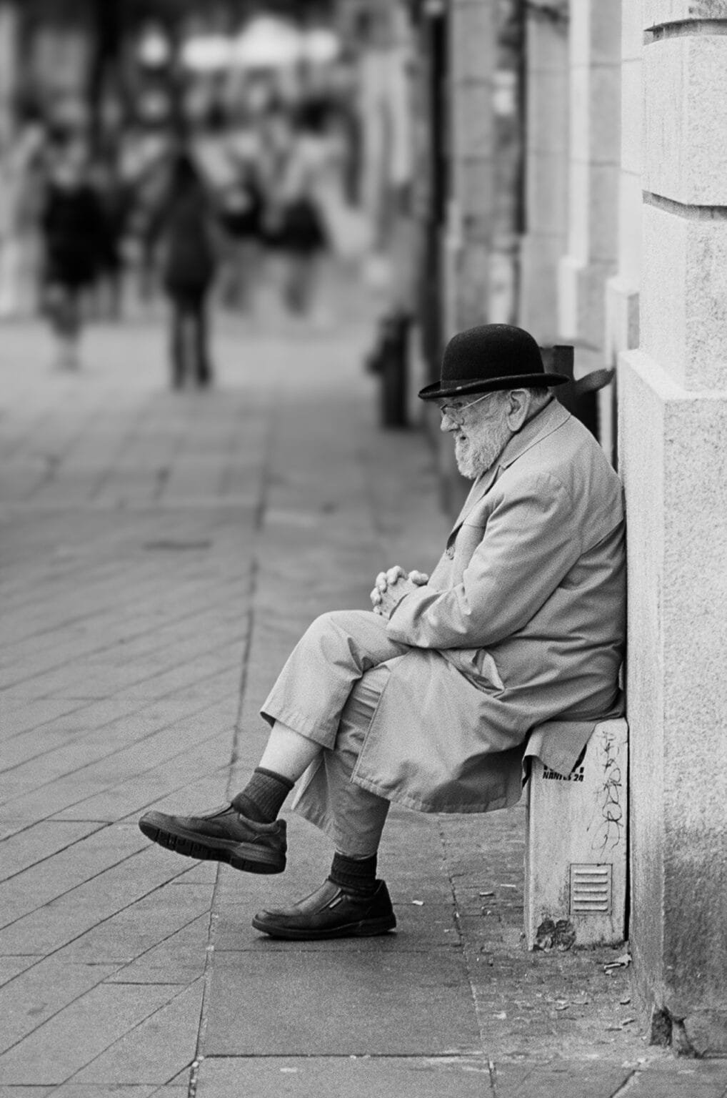Homme âgé assis sur un muret dans une rue de Nantes, portant un chapeau melon et un manteau.
