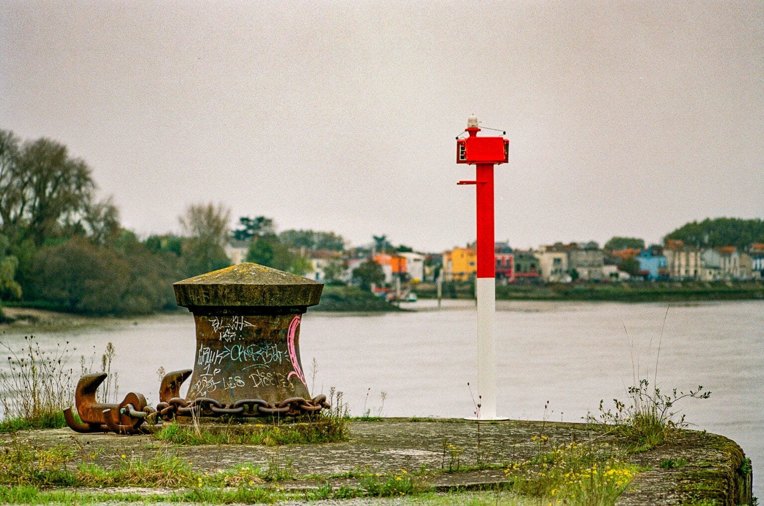 Bollard de quai rouillé avec une ancre, à Trentemoult, sur les rives de la Loire à Nantes, avec en arrière-plan les maisons colorées du village.