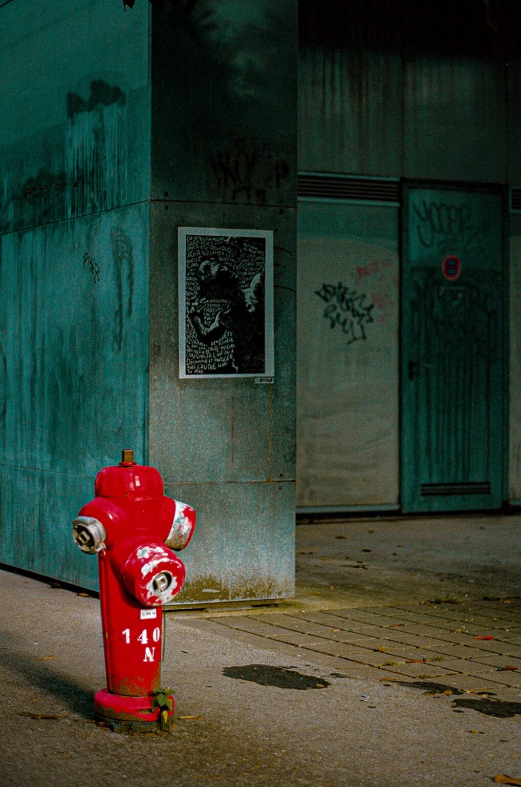 Borne incendie rouge usée sur le trottoir, avec en arrière-plan un mur tagué et une affiche collée, à Nantes.
