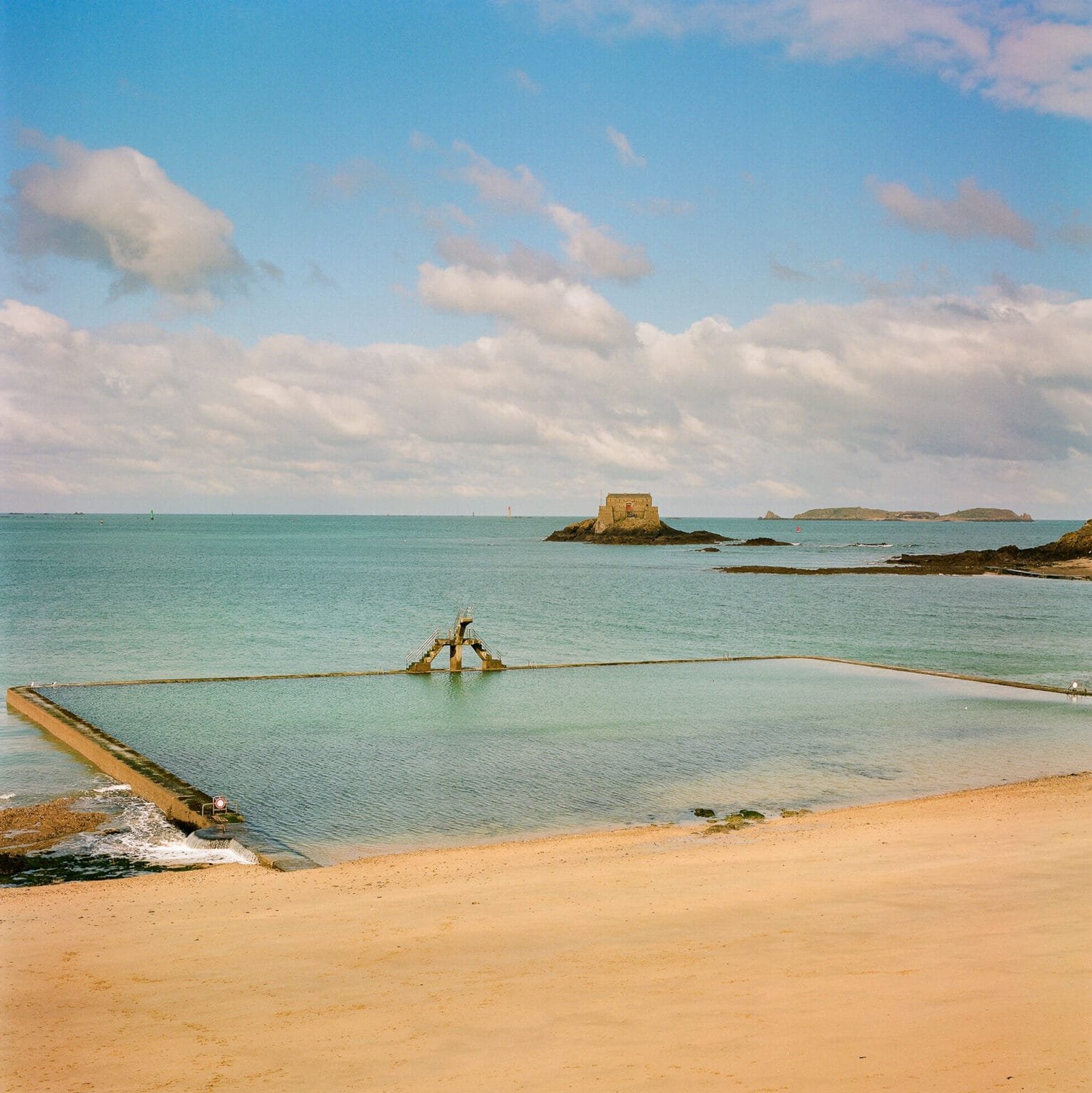 Vue sur la piscine d’eau de mer de Saint-Malo à marée basse, avec le fort visible au loin sous un ciel légèrement nuageux.