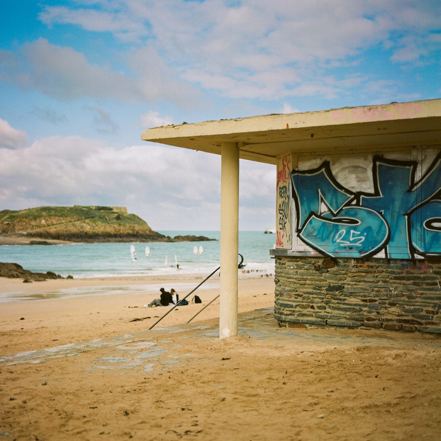 Abri de plage tagué sur la plage de Saint-Malo, avec des voiliers au loin sur la mer turquoise.