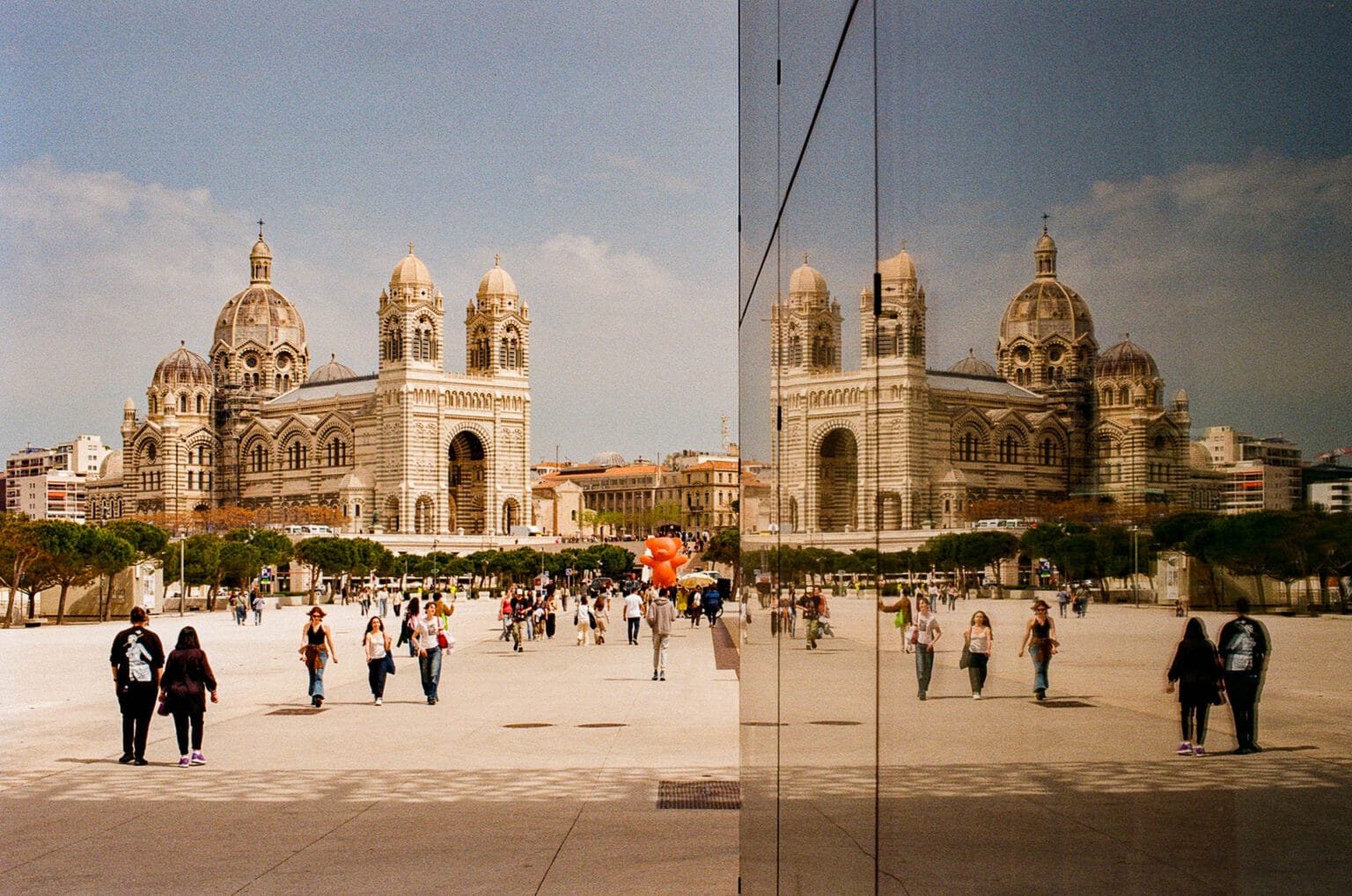 Reflet de la cathédrale de la Major à Marseille dans la façade vitrée du Mucem, avec des passants sur l’esplanade.