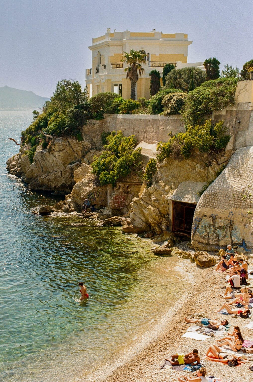 Petite plage rocheuse à Marseille, avec des baigneurs et une villa jaune surplombant la mer Méditerranée.