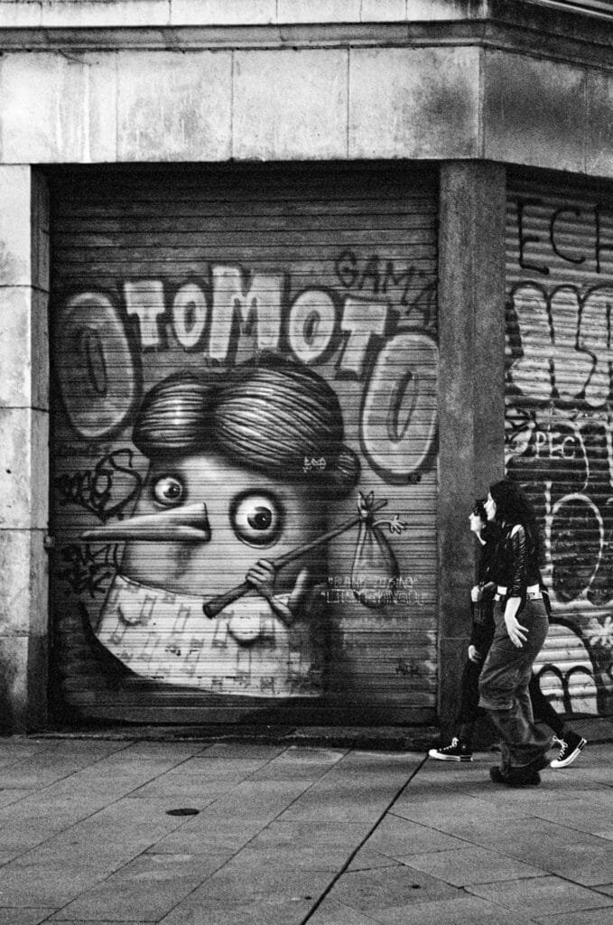 Scène urbaine en noir et blanc capturée à Nantes, avec un graffiti Otomoto et deux passants, prise avec un Rollei 35 S sur pellicule Ilford HP5.