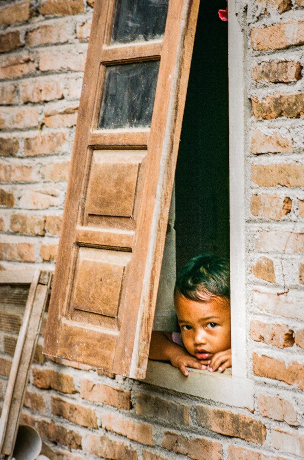 Enfant accoudé à une fenêtre entrouverte dans un mur en briques, regardant dehors.