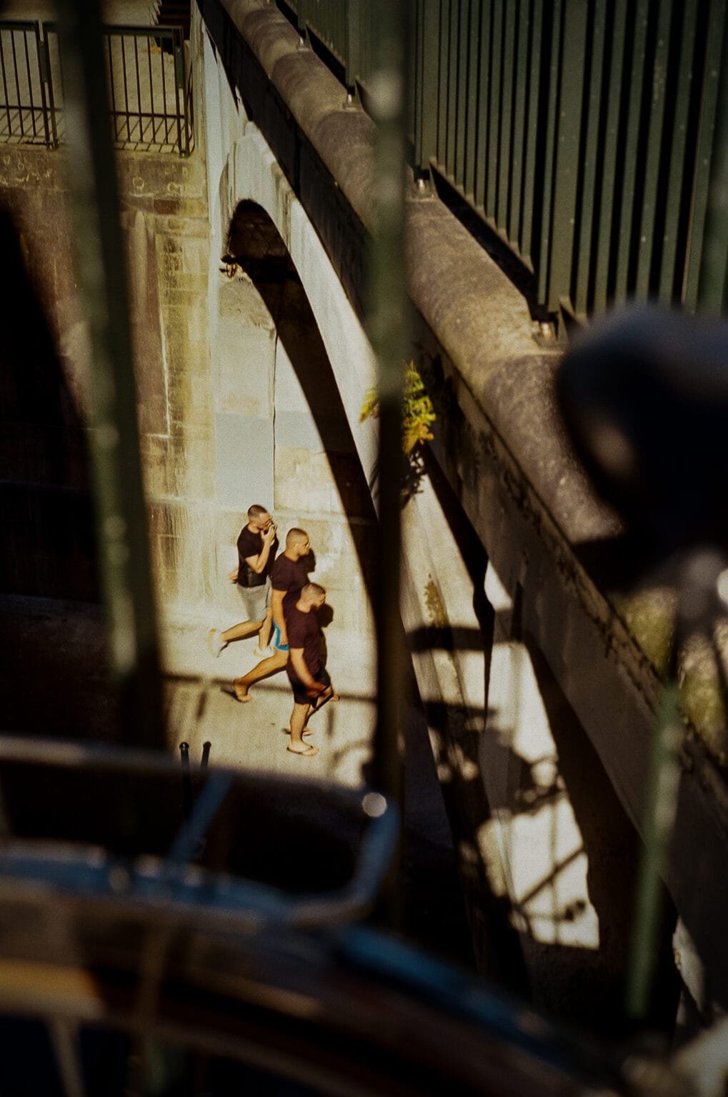 Trois jeunes hommes marchant pieds nus dans une contre-allée ensoleillée du centre de Nantes, photographiés en couleur à travers une rambarde.