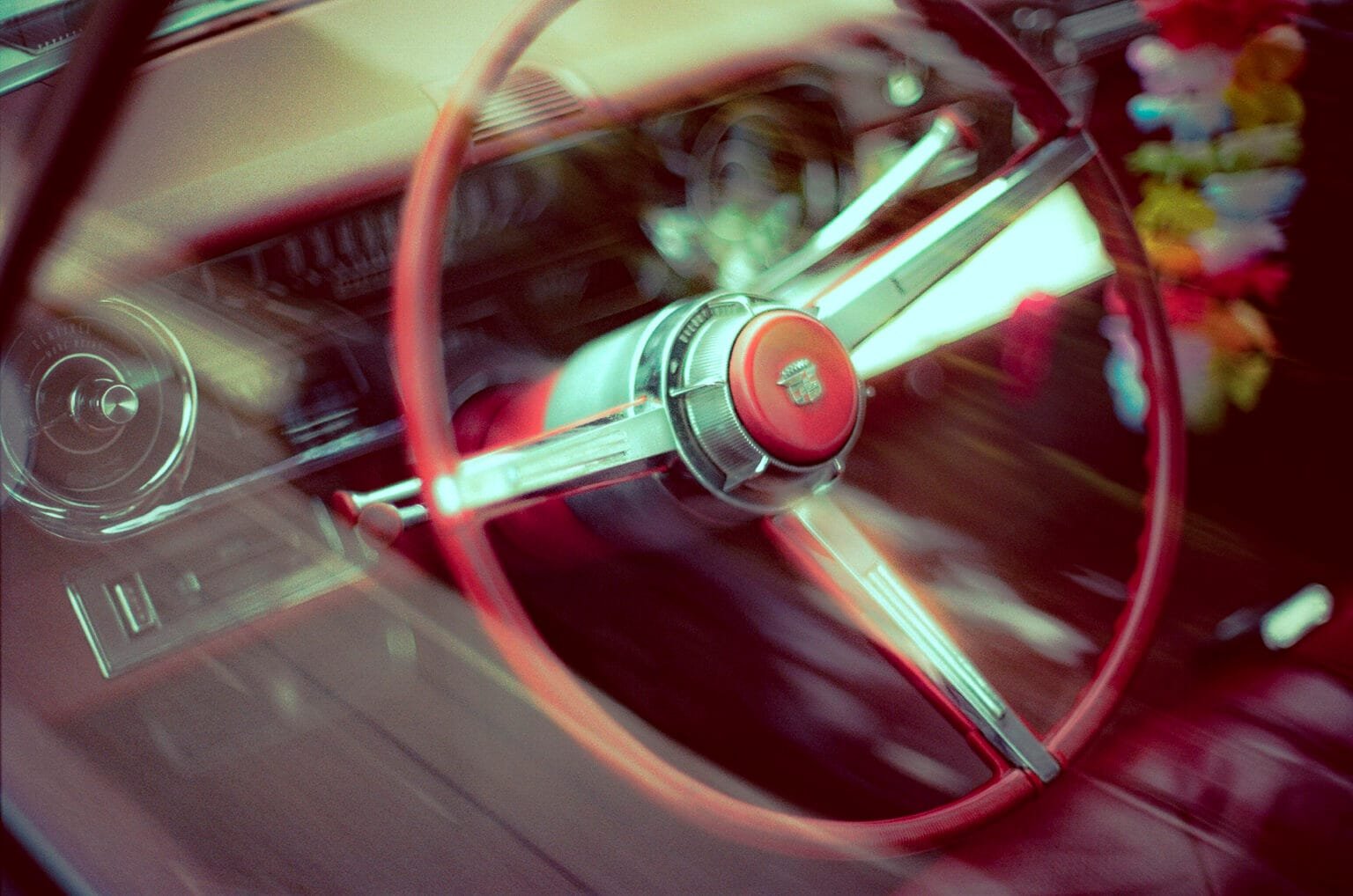 Volant rouge et chrome d’une Cadillac ancienne vu à travers la vitre, avec un collier de fleurs suspendu