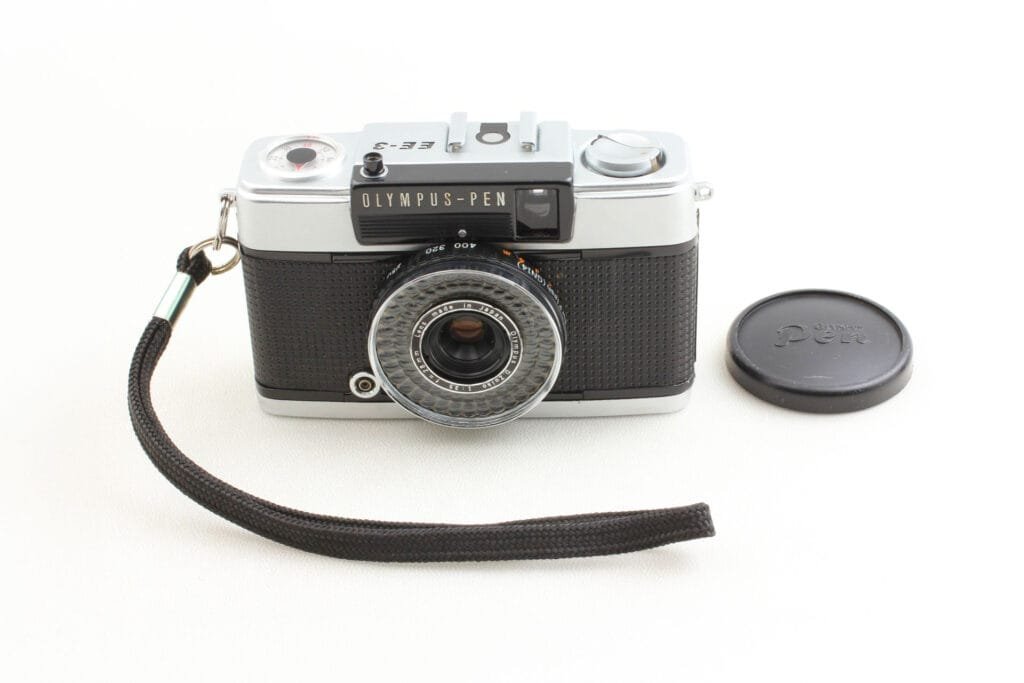 Olympus Pen EE-3 posé à plat, avec dragonne noire et bouchon d’objectif sur fond clair.