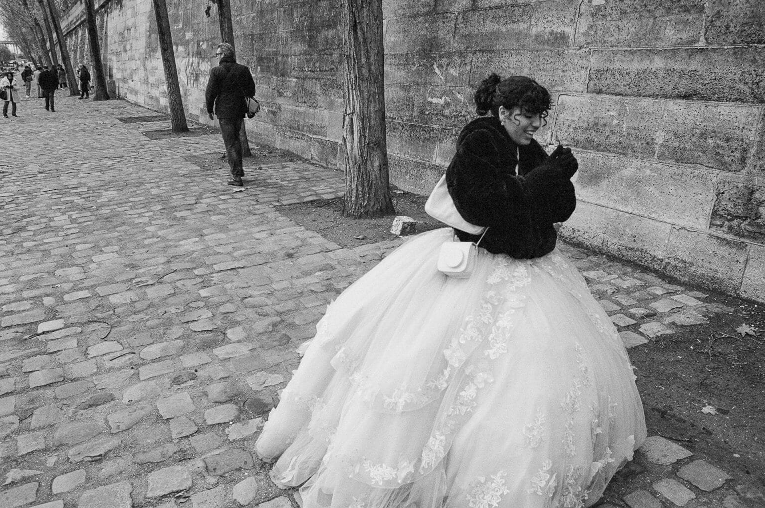 Jeune femme en robe de mariée et manteau noir marchant sur les quais pavés de Paris, en hiver