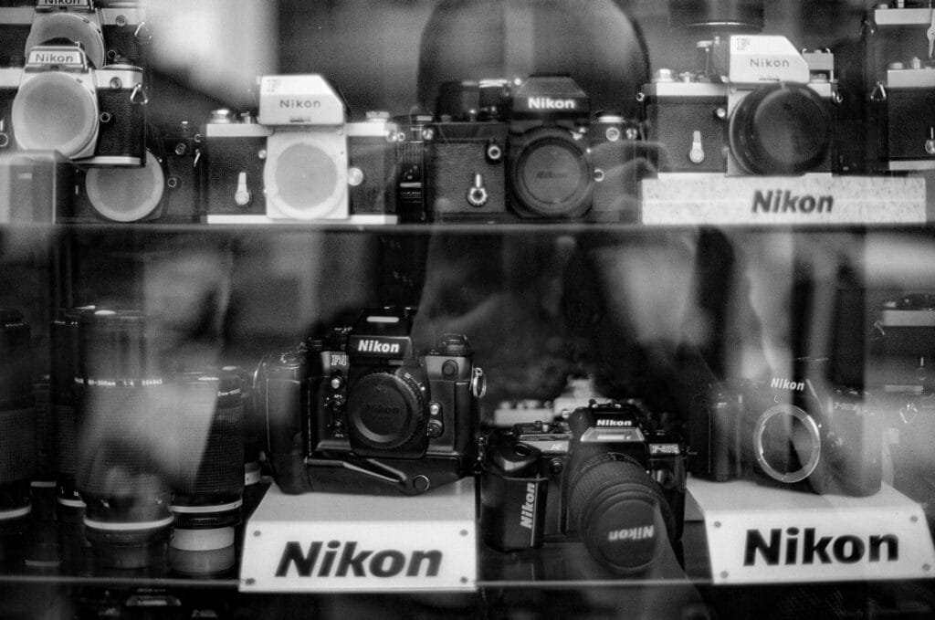 Vitrine de magasin remplie d’appareils photo Nikon vintage et objectifs, photographiée en noir et blanc à travers la vitre.