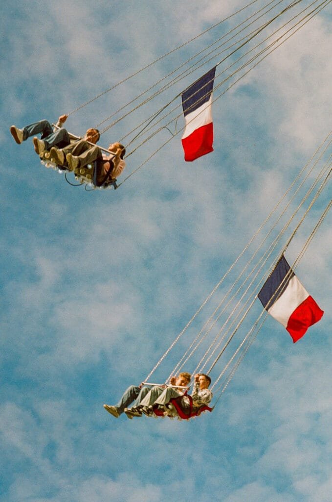 Des adolescents sur des balançoires volantes sous un ciel bleu à la fête foraine de Nantes, avec des drapeaux français flottant au vent.