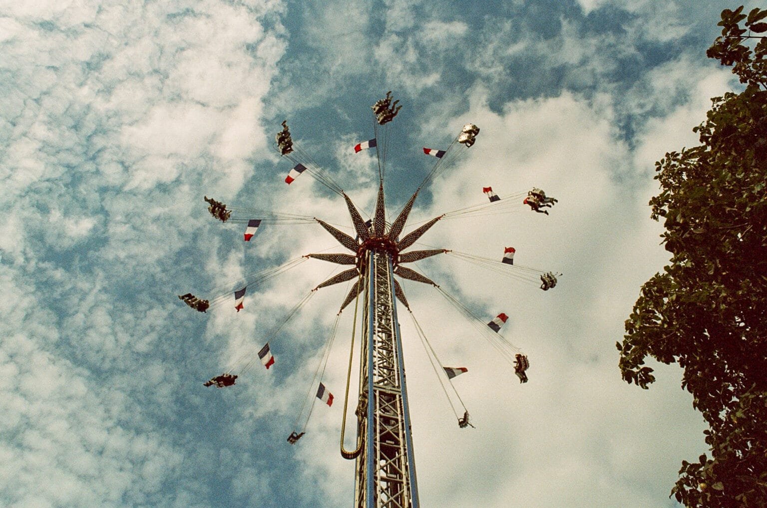 Vue en contre-plongée d’une attraction de chaises volantes à la fête foraine de Nantes, avec des drapeaux français flottant sous un ciel nuageux.
