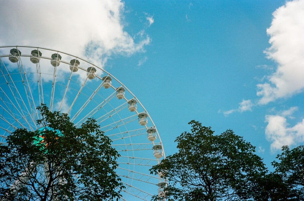 Vue partielle de la grande roue de Nantes en arrière-plan, avec des arbres au premier plan, sous un ciel bleu. Photographie en pellicule couleur avec un Olympus AF-1.