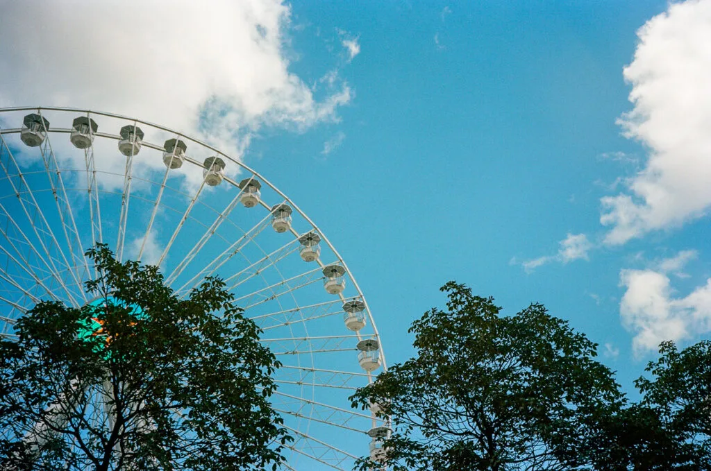 Vue partielle de la grande roue de Nantes en arrière-plan, avec des arbres au premier plan, sous un ciel bleu. Photographie en pellicule couleur avec un Olympus AF-1.