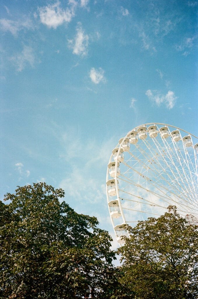 Vue partielle d’une grande roue blanche dépassant des arbres sous un ciel bleu à Nantes, prise avec un Olympus AF-1 sur pellicule Kodak Portra 160.