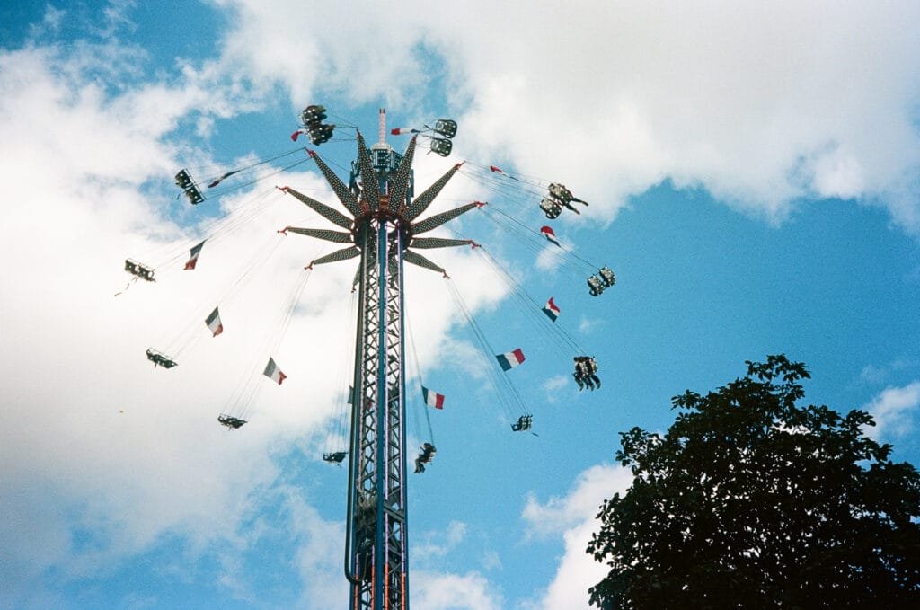 Manège à chaises volantes tournant dans le ciel de Nantes, avec des drapeaux français et un arbre en contrebas. Photo prise avec un Olympus AF-1 et une pellicule Portra 160.