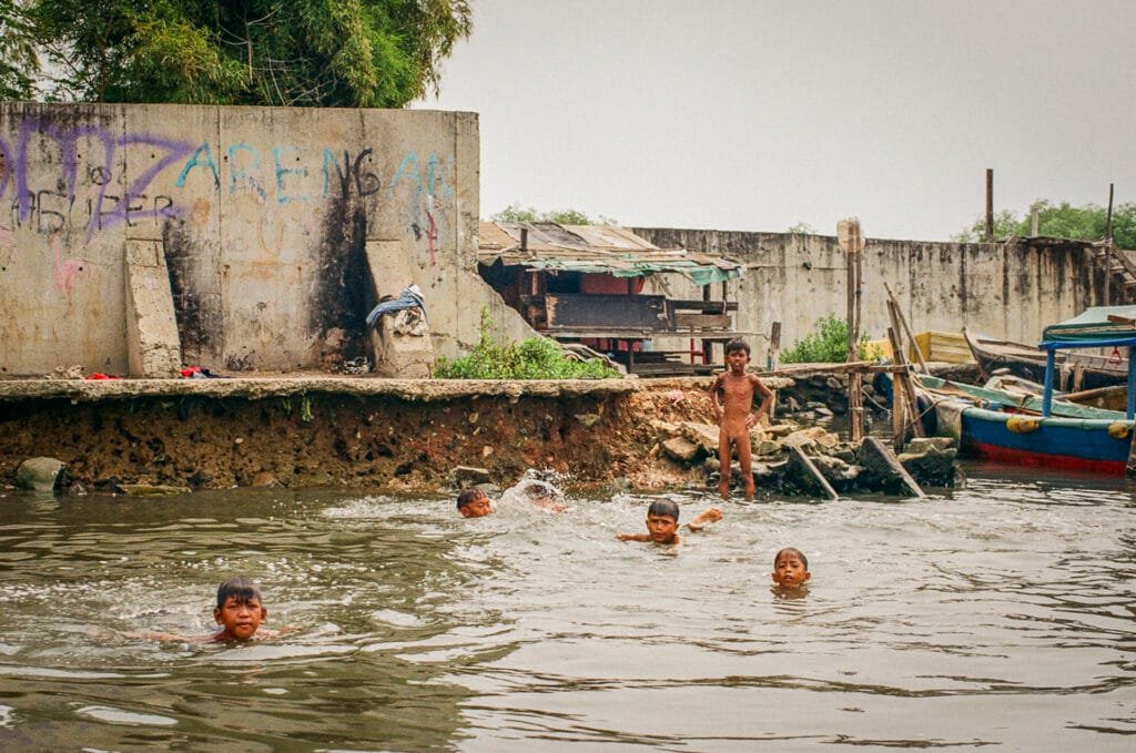 Enfants jouant et nageant dans l’eau du port de Jakarta, près de bateaux colorés et de baraques en bois.