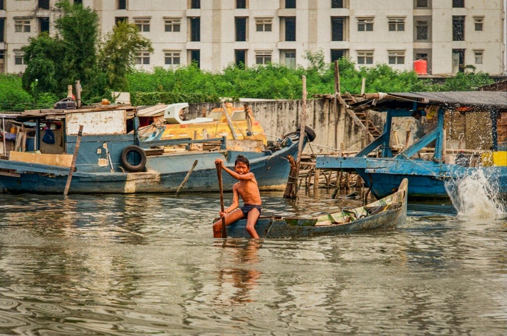 Jeune garçon maniant une pagaie dans une petite barque rouillée, entouré de bateaux de pêche, dans le port de Jakarta.