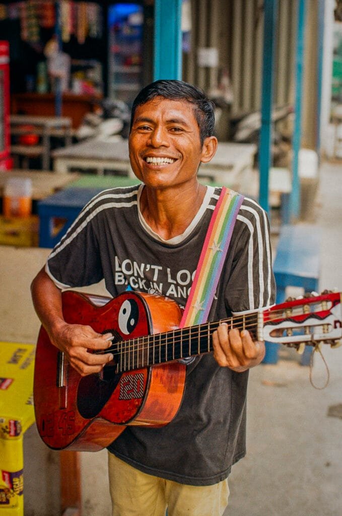 Homme souriant jouant de la guitare avec une sangle arc-en-ciel dans une rue du port de Jakarta.