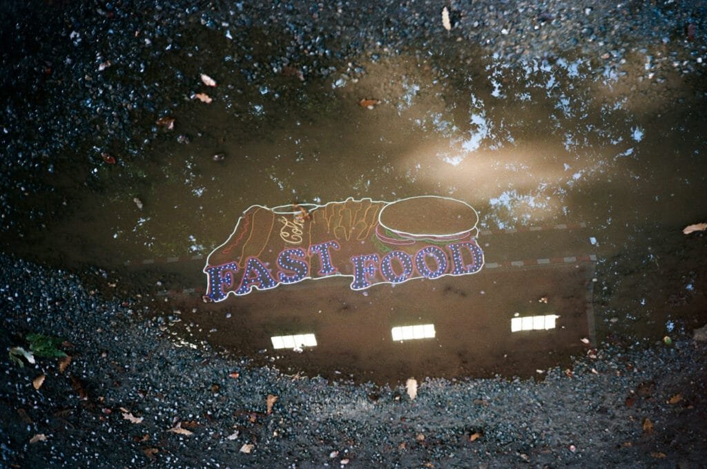 Reflet dans une flaque d’un néon « Fast Food » avec hot-dog, frites et burger à Nantes, photographié avec un Olympus AF-1 sur pellicule Kodak Portra 160.
