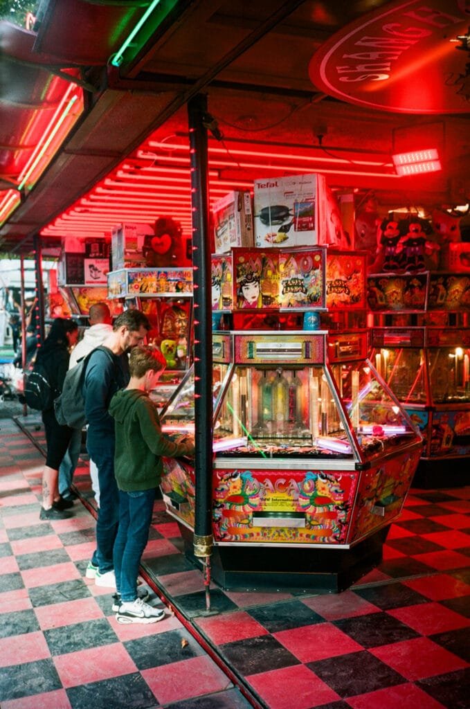 Un père et son fils jouent à une machine de type pousse-pièces dans une salle de jeux foraine éclairée de néons rouges.