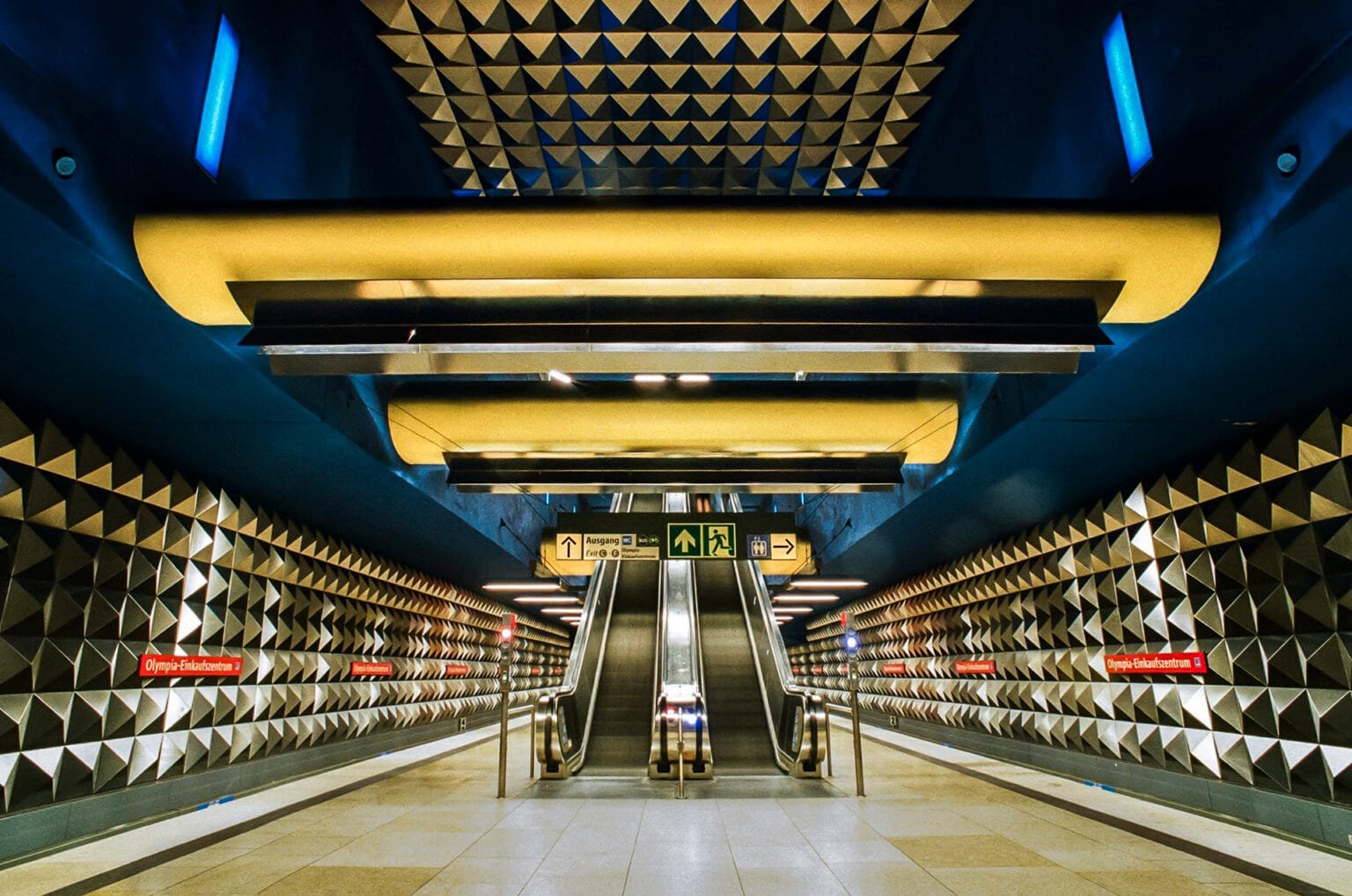 Vue symétrique des escalators de la station de métro Olympia-Einkaufszentrum à Munich, avec murs à motifs géométriques.