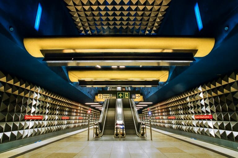 Vue symétrique des escalators de la station de métro Olympia-Einkaufszentrum à Munich, avec murs à motifs géométriques.