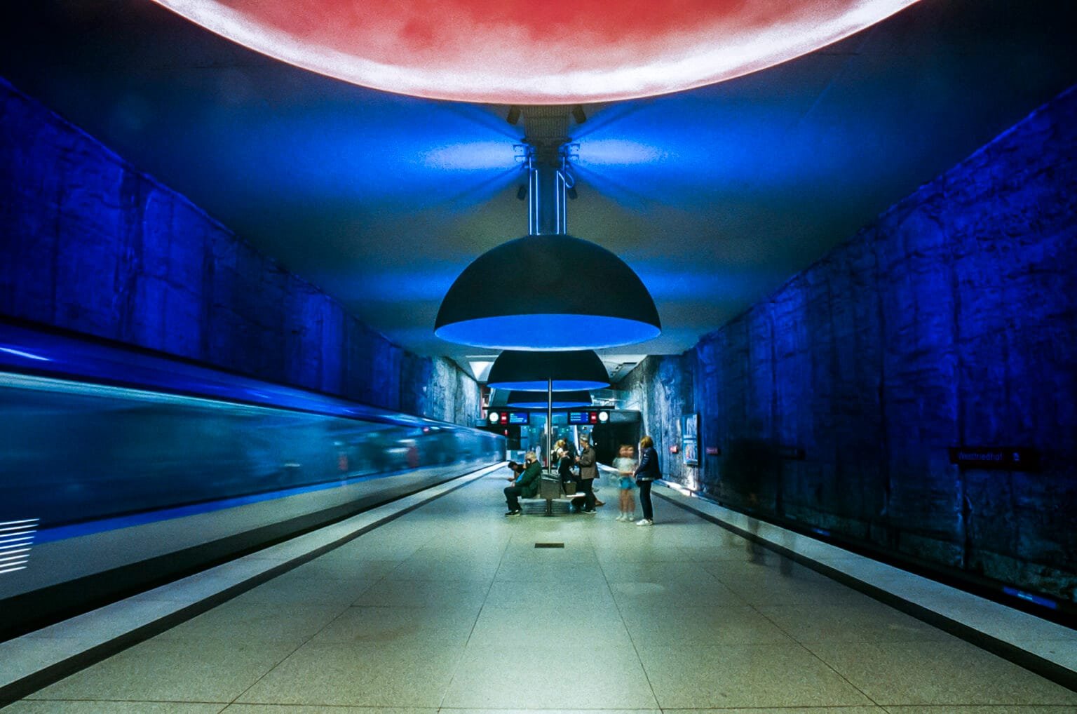 Station de métro Westfriedhof à Munich éclairée de lumières rouges et bleues, avec un train en mouvement.