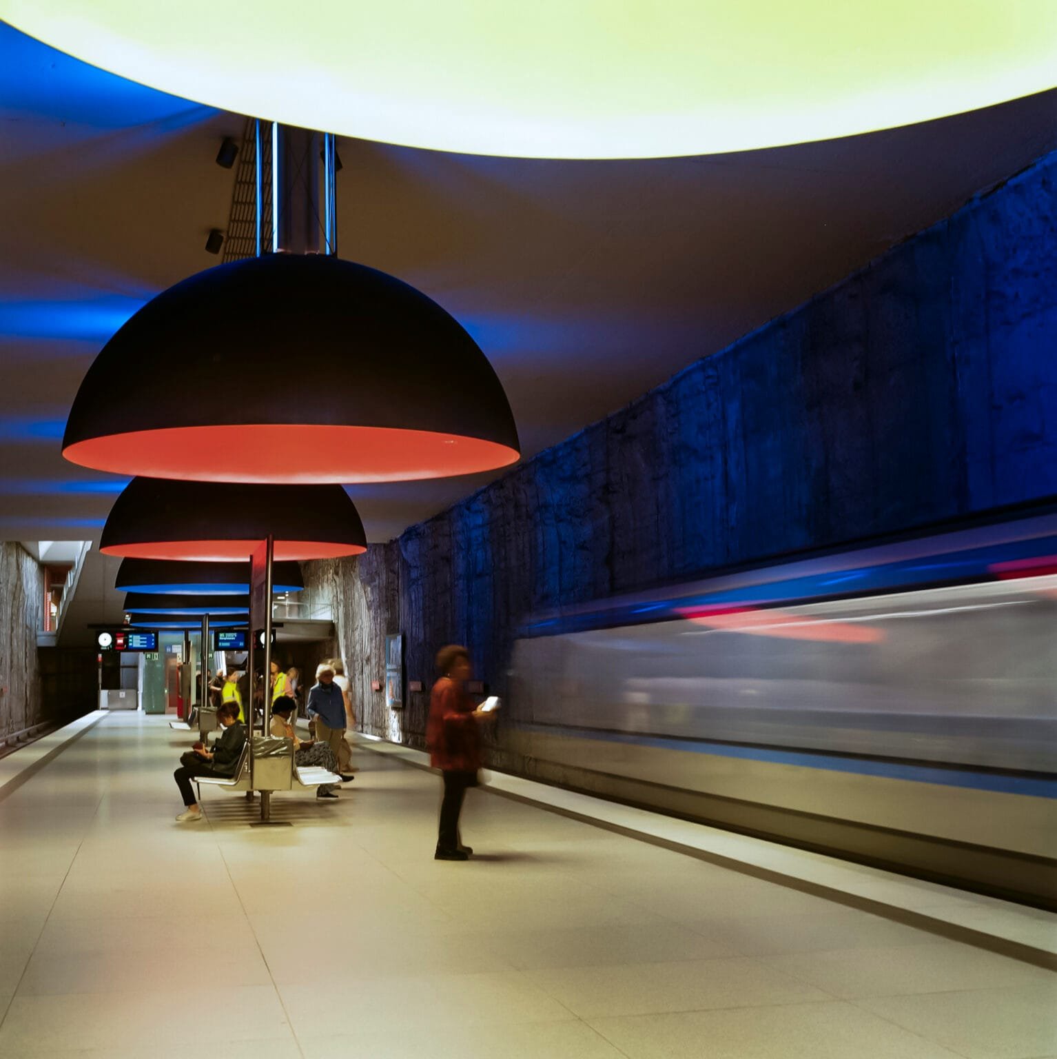 Train en mouvement dans la station de métro Westfriedhof à Munich, sous des suspensions colorées et un éclairage bleu.