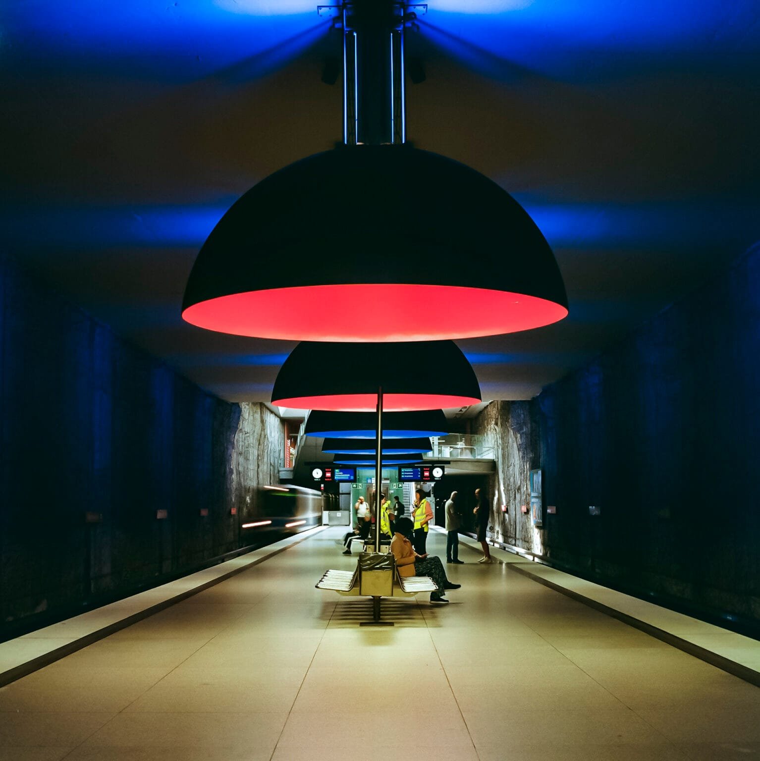 Vue centrale de la station de métro Westfriedhof à Munich, sous des suspensions rouges et un éclairage bleuté.