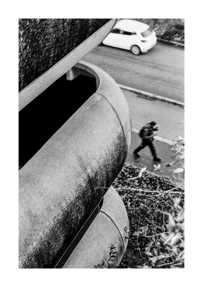 Vue oblique du parking Gare Nord de Nantes en béton, avec un passant flou et une voiture blanche en arrière-plan, photographié en noir et blanc.