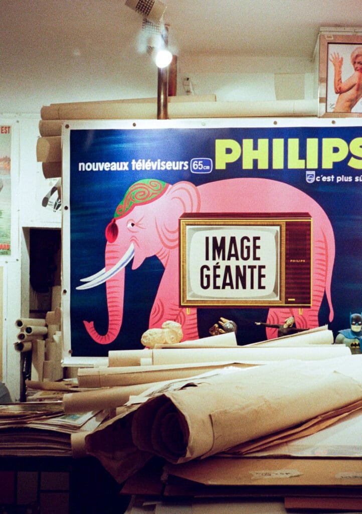 Affiche publicitaire vintage Philips avec un éléphant rose et un vieux téléviseur, photographiée dans un atelier encombré de rouleaux de papier.