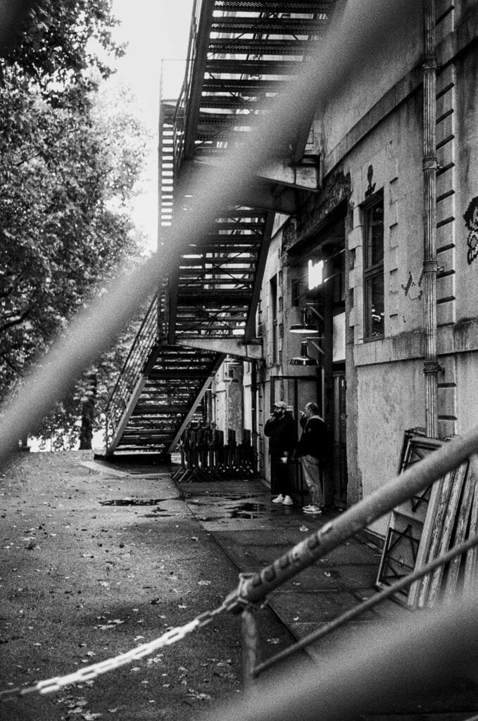 Vue en noir et blanc de l’extérieur du Lieu Unique à Nantes, avec deux personnes sous un escalier métallique, photographiées à travers une barrière.