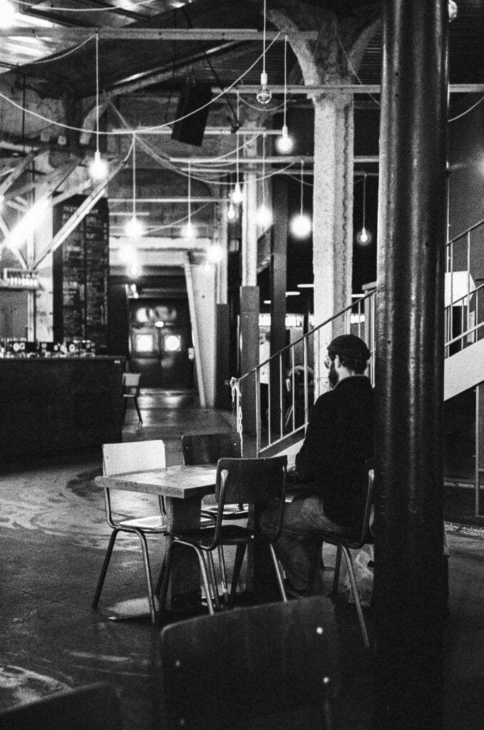 Homme assis seul à une table du bar du Lieu Unique à Nantes, dans une ambiance tamisée et industrielle, photographié en noir et blanc.