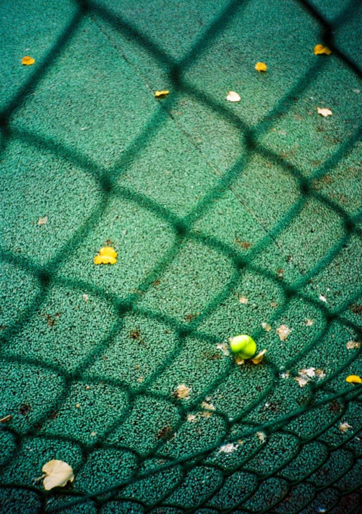 Balle de tennis sur un terrain synthétique vert, vue à travers un grillage, au parc du Luxembourg à Paris. Pellicule Portra 160, Canon Dial 35-2.