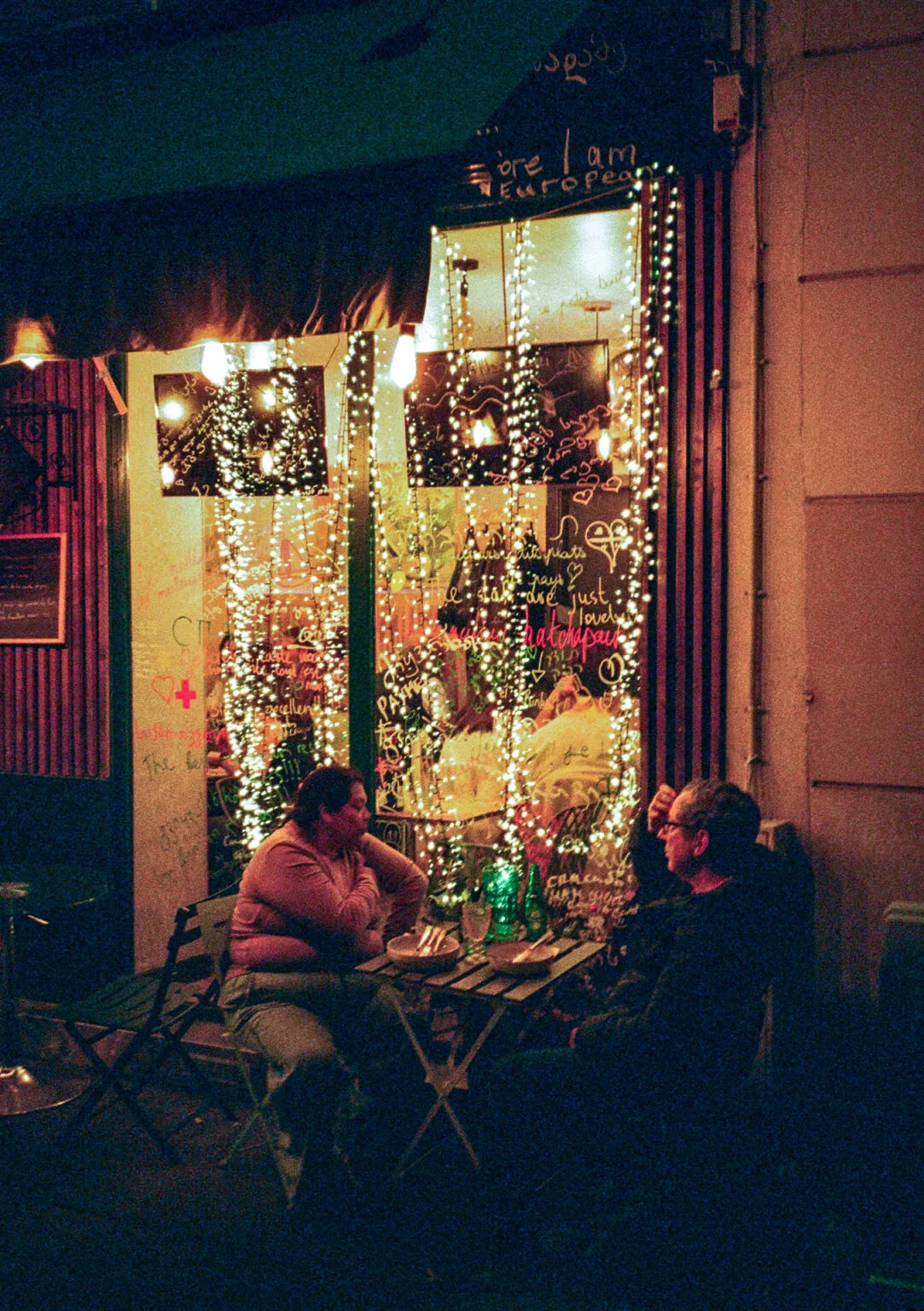 Deux personnes discutent en terrasse le soir, devant une vitrine couverte de guirlandes et d’inscriptions colorées.