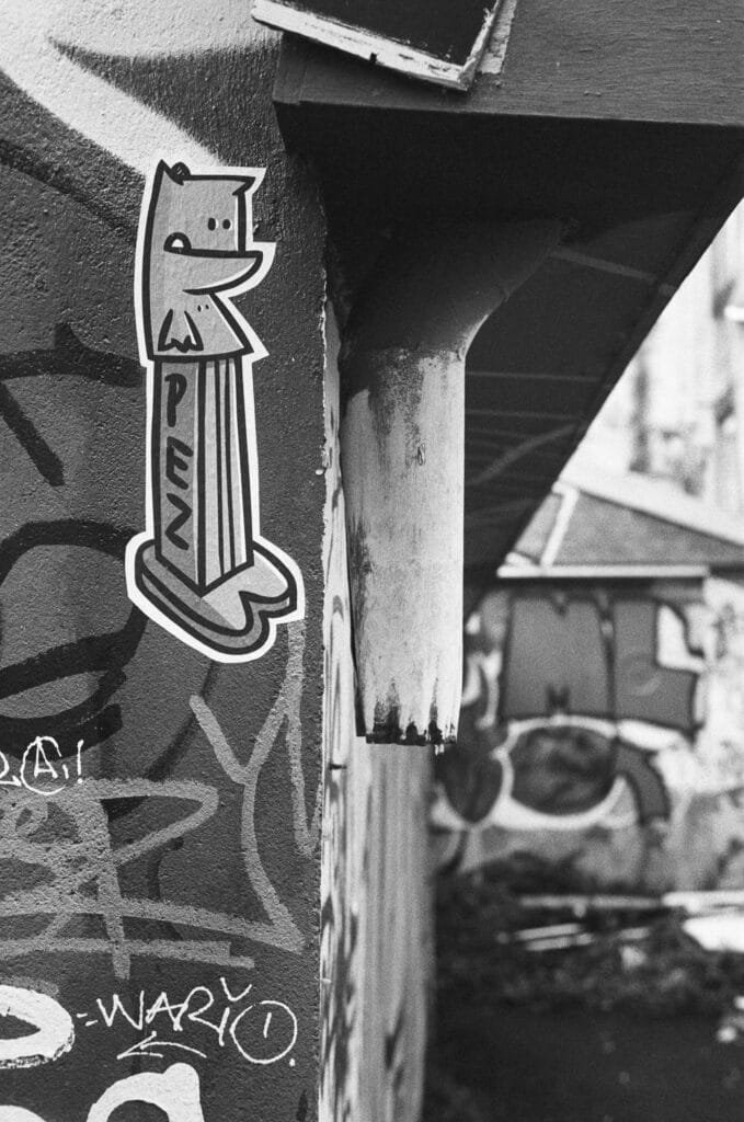 Sticker PEZ en forme de chat collé sur un mur tagué, avec une gouttière rouillée et des graffitis en arrière-plan.