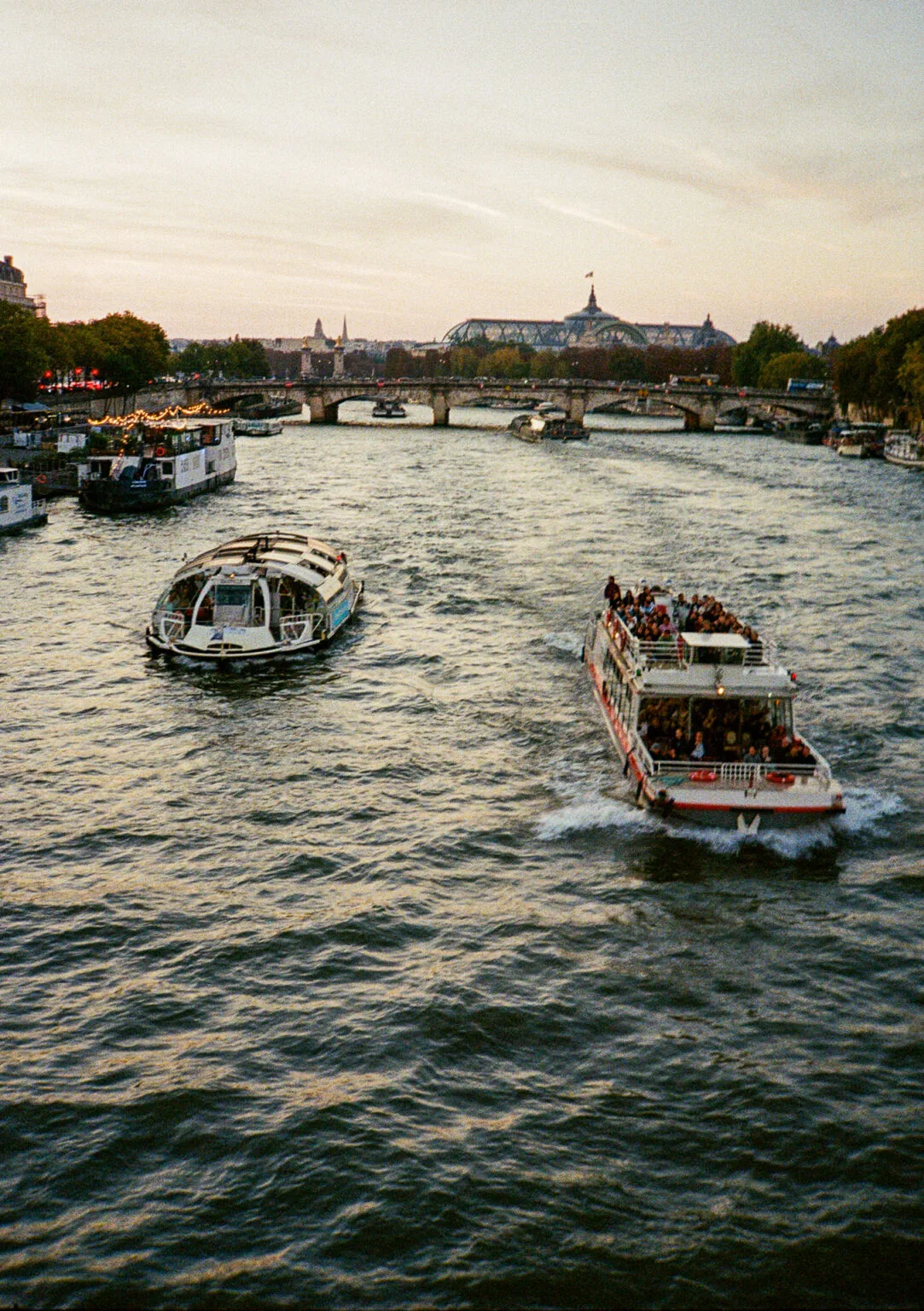 Vue sur la Seine à Paris au coucher du soleil avec deux bateaux-mouches en mouvement, et le Grand Palais à l’arrière-plan.