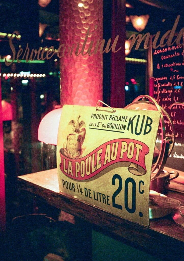 Affiche vintage “La poule au pot” dans la vitrine d’un restaurant parisien, éclairée par des lampes rouges et des néons.