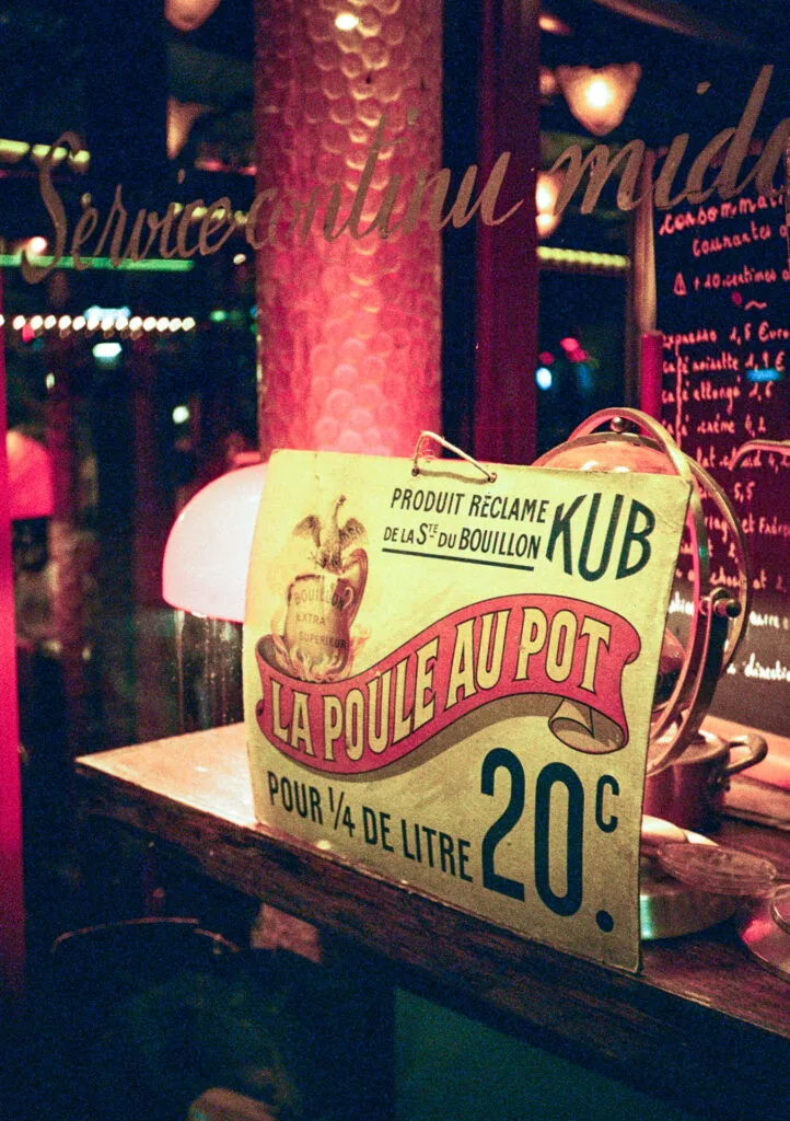 Affiche vintage “La poule au pot” dans la vitrine d’un restaurant parisien, éclairée par des lampes rouges et des néons.