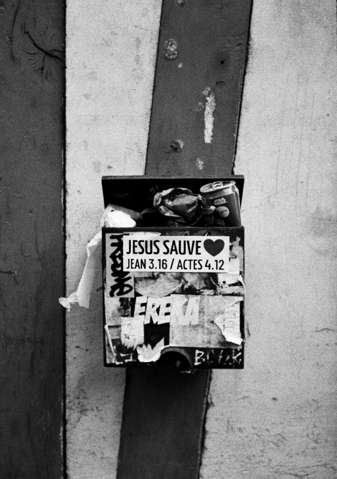 Boîte saturée de détritus recouverte d’autocollants religieux et de graffitis, photographiée en noir et blanc à Nantes.