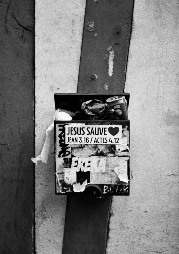Boîte saturée de détritus recouverte d’autocollants religieux et de graffitis, photographiée en noir et blanc à Nantes.